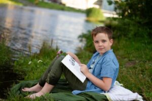 Child sitting barefoot near a river, reading a book on a summer day.